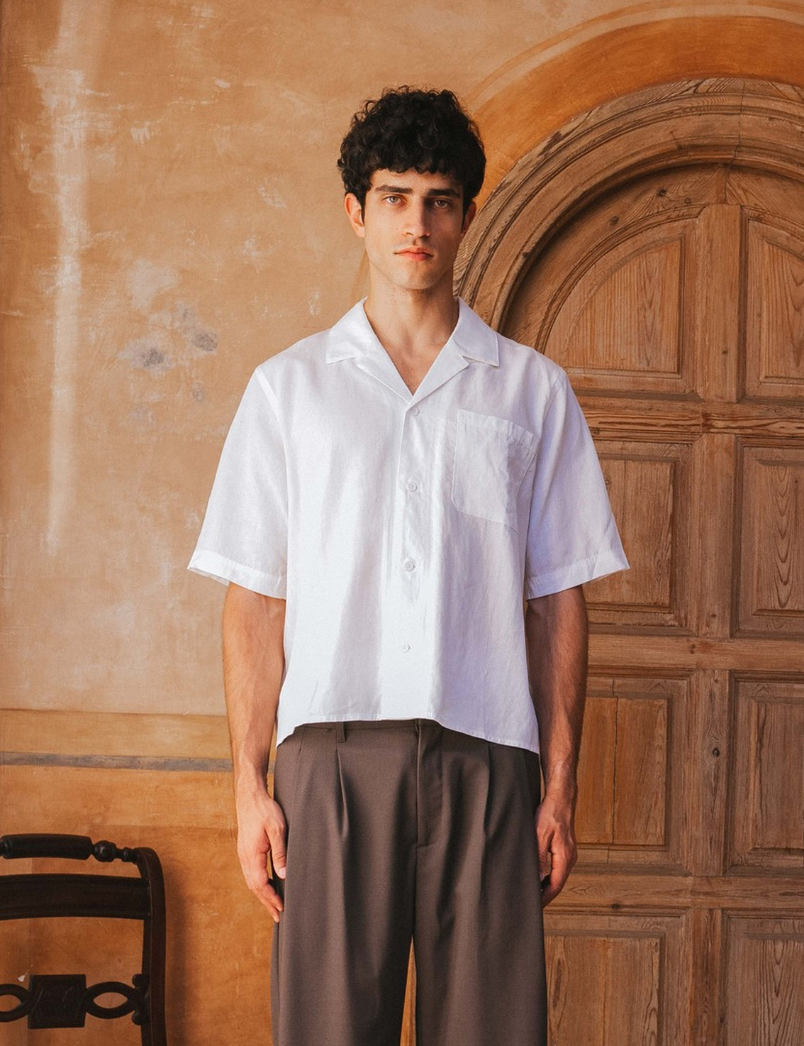 Man wearing a white shirt and brown pants standing in front of a wooden door.